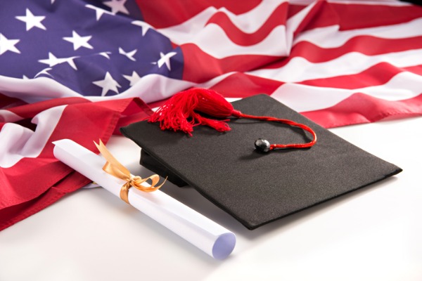 closeup-view-of-graduation-mortarboard-diploma-and-us-flag-on-white-picture-id6654347222